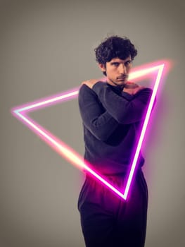 A man stands inside of a neon triangle sign, illuminated against a dark background. The man appears to be observing the vibrant color and geometric shape of the neon triangle.