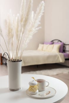Sweet breakfast in the bedroom. A vase with decorative flowers, a cream cake and a cup of tea on a white table against the background of the bed in the bedroom.