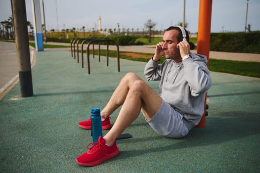 Young athletic man putting on wireless headphones, resting after workout, sitting on the sports ground. Active exhausted sportsman relaxing after a bodyweight sports training outdoors on a sunny day