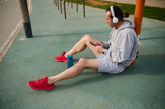 Young active athletic man in wireless headphones, checking mobile app on his smartphone while resting after workout the urban sports ground. Sportsman using mobile phone while working out outdoors