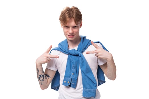 young well-groomed cute caucasian male student with golden red hair with a tattoo on his arms is dressed in a white t-shirt.