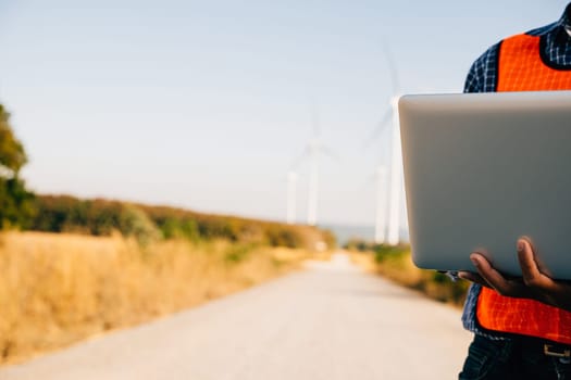Engineer laptop in hand assesses wind turbines. Service technician ensures efficiency. Focused on innovation quality and landscape development in windmill service.