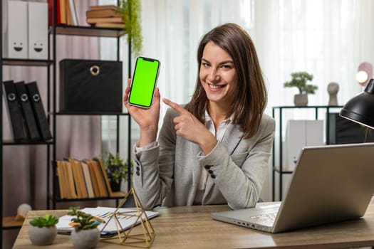 Caucasian young businesswoman holding smartphone with green screen chroma key mock up recommend good application promotional sale offer. Freelancer woman girl at home office desk looking at camera.