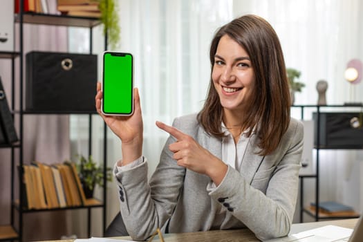 Happy smiling young businesswoman holding smartphone with green screen chroma key mock up recommend good application promotional sale offer. Freelancer woman girl at home office desk looking at camera
