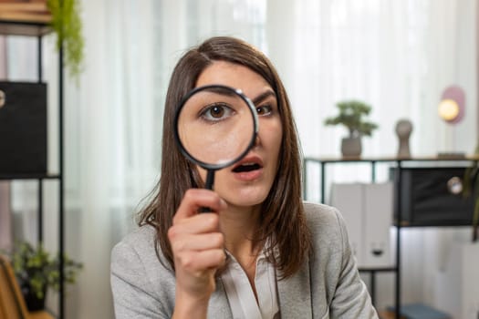 Investigator researcher businesswoman looking through magnifying glass with wow expression with big zoomed funny eye, searching, analyzing. Professional Caucasian freelancer girl looking at camera.
