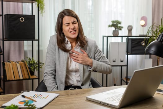 Stressed Caucasian young businesswoman using laptop and suffers from tension holding chest with heart pain attack risk of stroke symptom while working at home office desk. Girl freelancer at work.