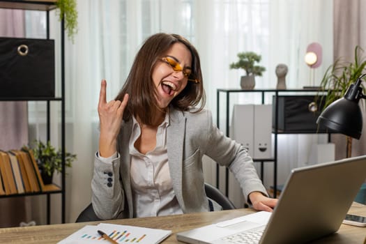 Happy excited Caucasian businesswoman working on laptop at home office desk putting on earbuds listening to favorite energetic disco music and dancing. Freelancer girl relaxing, taking a break.