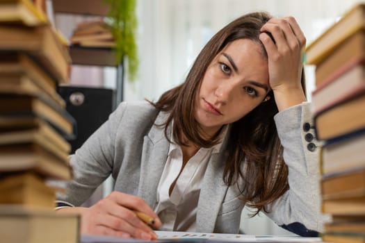 Exhausted Caucasian business woman looking at documents reports while working hard at home office desk. Stressed alone girl freelancer touching head in despair surrounded by stacks of books on table.