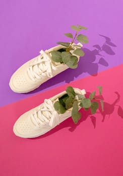 One modern fashionable white sneakers with eucalyptus branches in them lie on a lilac-pink background with hard shadows, flat lay close-up. Fashionable shoes concept.