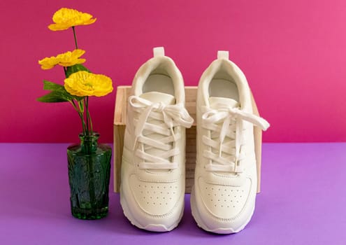 One pair of modern fashionable white sneakers rests on a wooden box with a nearby vase of yellow flowers on a lilac-pink background, close-up side view. Fashionable shoes concept.