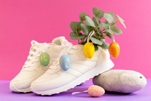 One pair of modern fashionable white sneakers with marble decorative, eucalyptus branches and Easter eggs rest on a stone on a lilac-pink background, close-up side view. Fashionable shoes concept.