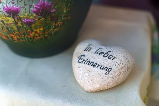 A heart-shaped rock lies next to a vase filled with colorful flowers, creating a sweet and natural arrangement. There is an inscription on the stone in German - In lovely memories