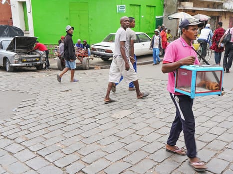 Antananarivo, Madagascar - April 24, 2019: Typical street scene in Antananarivo - some Malagasy people sitting on stone ground porch, man fixing his car near, other is selling freshly baked pastry