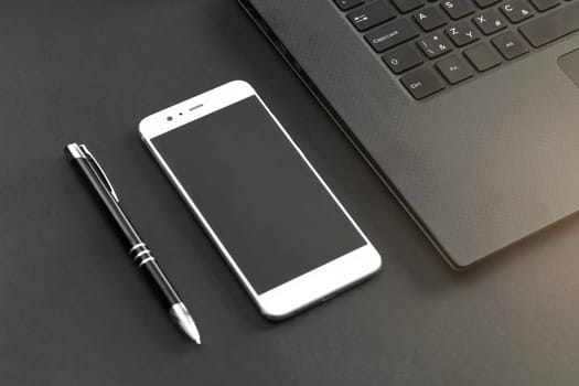 Pen, mobile phone, and laptop keyboard on gray / black desk - view from above