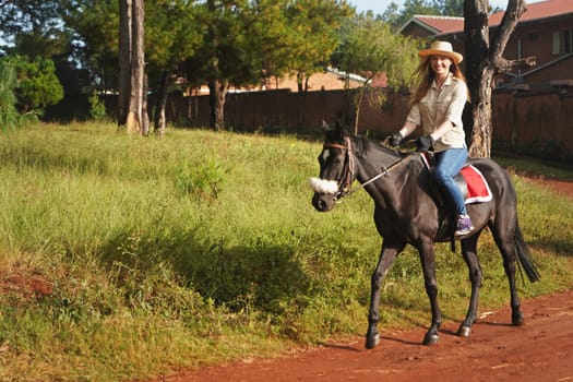 Young woman in shirt and straw hat, riding black horse in the park, blurred background with houses and trees