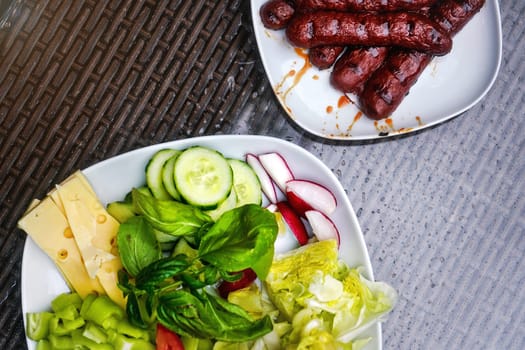 Two white plates, one with grilled bratwursts / frankfurters, other with salad - cucumbers, radish, basil, pepper and cheese. View from above