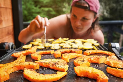 Butternut squash pieces grilled on electric grill, focus on bright orange vegetables seasoned with spice, blurred young woman in background