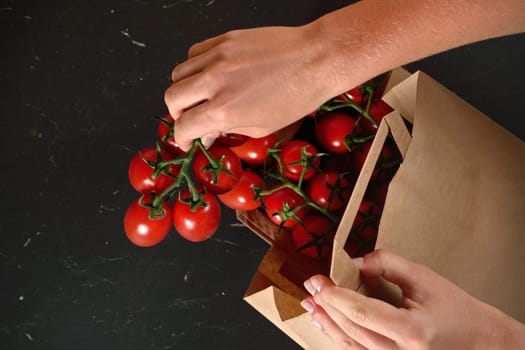 Taking out red cherry tomatoes with green leaves from brown paper bag over dark marble like board, detail on woman hand holding fruits