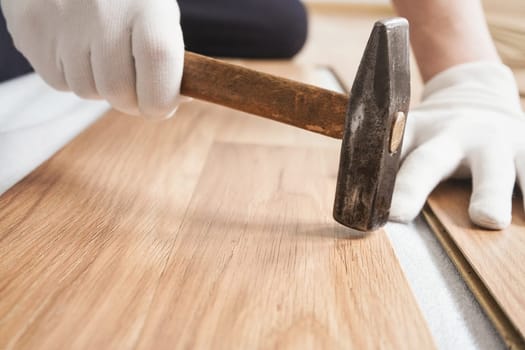 Installing laminated floor, detail on man hands in white gloves, holding hammer over wooden tile, white base layer under