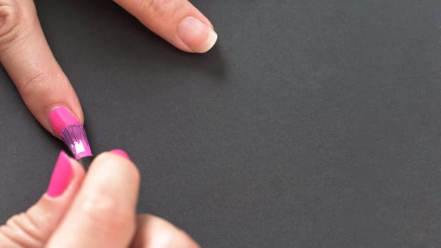 Applying pink nail polish - closeup photo of woman hands, little brush and fingernails on gray / black background, space for text right side