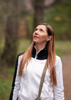 Young woman wearing white sport jacket walks in forest, looks up to sky, blurred trees behind her