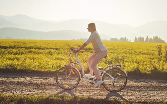 Young woman rides electric bike over dusty country road, strong afternoon sun backlight in background shines on yellow flowers field, view from side