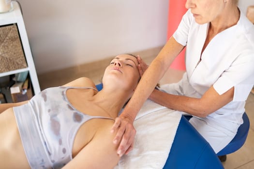 Crop physiotherapist in white uniform holding shoulder and stretching neck of patient lying in bed and closing eyes during massage procedure