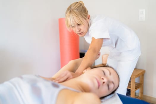 Focused female physiotherapist in white uniform massaging armpit of woman lying on couch during osteopathy session