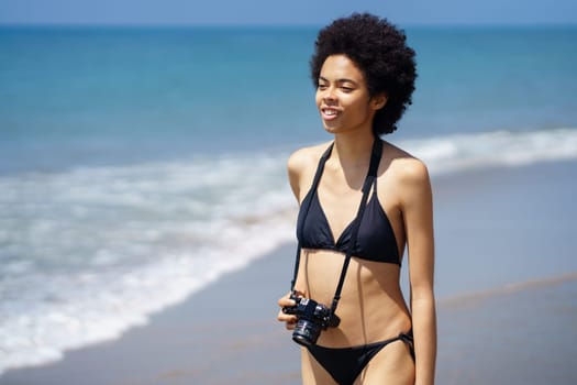 Optimistic African American female in swimwear with professional photo camera standing on wet seashore near waving sea in tropical resort in sunny weather