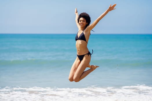 Full body of smiling carefree African American female traveler in swimwear looking at camera while jumping with raised arms on coast near sea