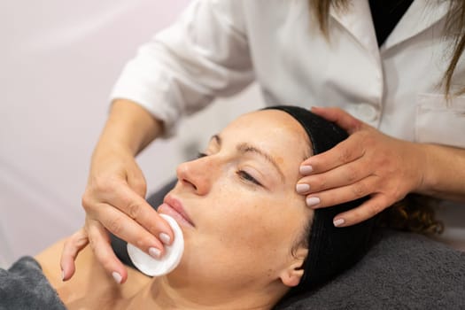 Crop cosmetician cleaning face of young female client with cotton pad during skincare treatment in modern beauty salon