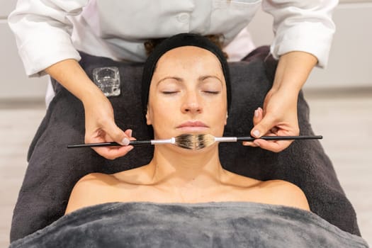 From above crop view of cosmetician applying cleansing oil with brush on face of woman lying face up eyes closed, and covered with gray towel during makeup session in beauty salon