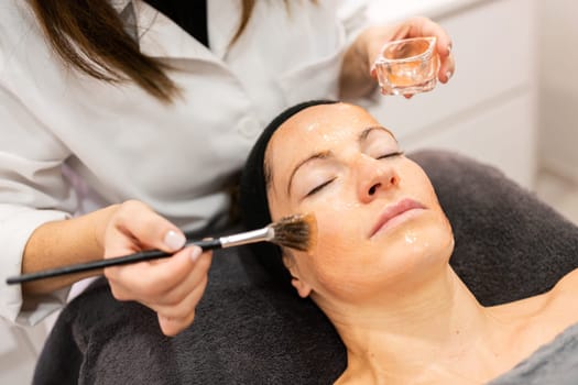 Unrecognizable cosmetologist in white uniform using brush and applying cosmetic product on face of female customer during facial beauty procedure in salon