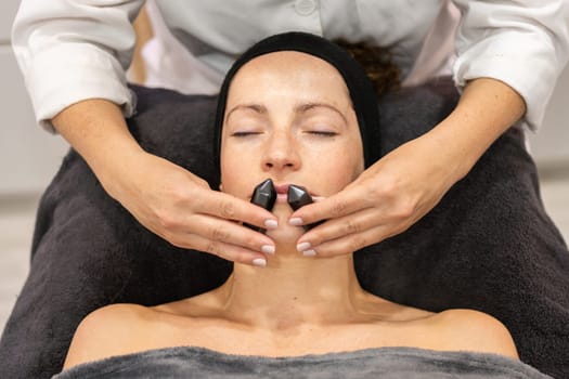 From above of crop anonymous cosmetician massaging face of relaxed woman with stones during beauty treatment in salon