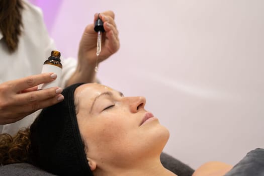 Crop beauty specialist applying serum on face of young female customer lying in modern salon during skin care routine