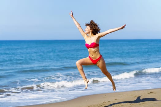 Smiling carefree young slim female redhead in pink bikini raising arms and jumping with legs in air while looking away, in daylight against blurred sea and blue sky