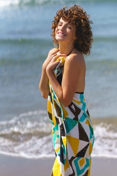 Side view of happy young sensual female with curly hair in colorful dress standing on seashore and looking at camera