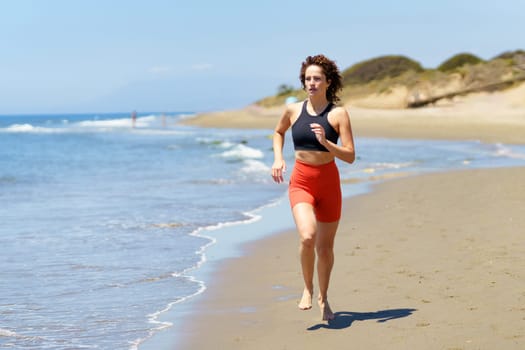 Full body of concentrated barefoot young female in sportswear warming up and looking away, while jogging on sandy beach near waving sea against blurred blue sky and grassy sand dunes