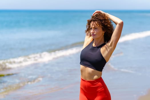 Thoughtful young female athlete in sportswear standing near waving sea and stretching arm behind back while looking away