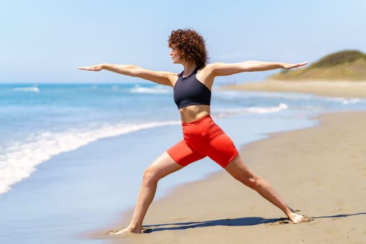 Full body side view of curly haired sportswoman practicing Warrior pose on sunny day near sea