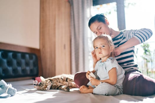 Mom combs a little girl sitting behind her on the bed near a lying cat. High quality photo