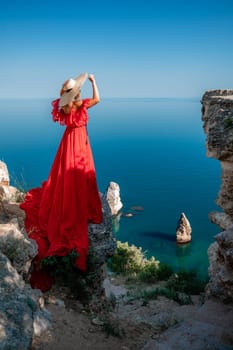 Red dress sea woman. Happy woman with flowing hair in a long flowing red dress stands on a rock near the sea. Travel concept, photo session at sea