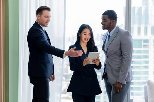 Diverse smart businesspeople standing near window with skyscraper while looking at business data analysis. Group of business team using tablet represent marketing plan, strategy, idea. Ornamented..