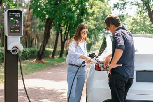 Lovely young couple wearing sun glasses recharging battery for electric car during road trip travel EV car in natural forest or national park. Eco friendly travel during vacation and holiday. Exalt