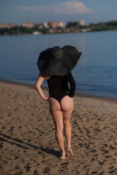 Rear view of woman in large straw hat and black swimsuit posing on the beach