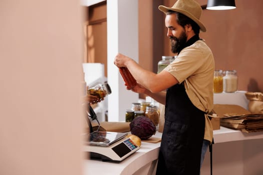 Caucasian male shopkeeper, who wears a stylish hat and black apron, stands behind the checkout counter holding a jar filled with pasta sauce. Vendor analyzing glass container at cashier desk.