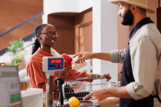 Black woman at checkout counter with a shopping basket filled with locally grown food products for vendor to weigh. Male cashier assisting african american client with her groceries.