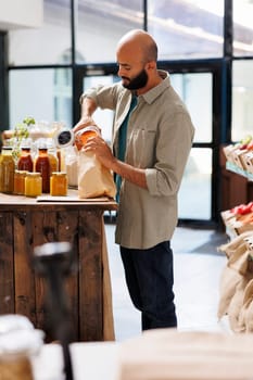 Middle Eastern man filling up a brown paper bag with orange lentils. Young man pouring grains into bag, buying healthy goods from reusable containers with organic bulk products.