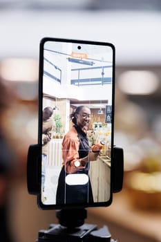 African American store owner uses cell phone and selfie stick for vlogging, promoting sustainable, healthy and natural items. Organic, fresh food in glass jars, appealing to eco-conscious customers.