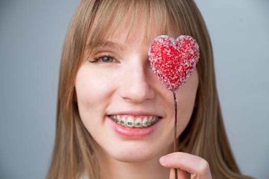 Cute woman with braces on her teeth holds a candy in the form of a heart on white background.